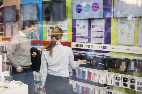 Verkäuferin, die einem Kunden beim Kauf einer Handyhülle hilft, gesehen durch Glas - MASF03419