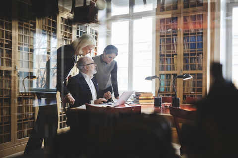 Älterer Anwalt diskutiert mit weiblichen Kollegen an einem Tisch in einer Bibliothek, durch Glas gesehen, lizenzfreies Stockfoto