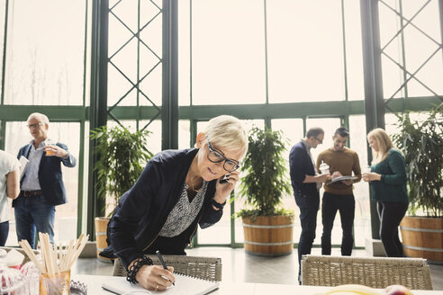 Reife Geschäftsfrau am Telefon, während sie in einen Notizblock mit Kollegen im Hintergrund im Büro schreibt - MASF03469