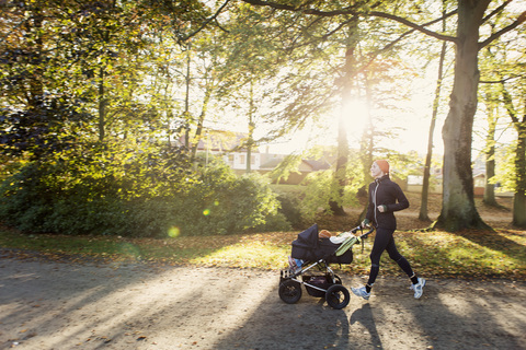Mutter joggt mit Kinderwagen auf der Straße gegen Bäume im Park, lizenzfreies Stockfoto
