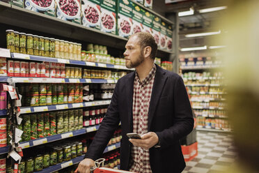 Männlicher Kunde hält Smartphone beim Einkaufen im Lebensmittelgeschäft - MASF03509