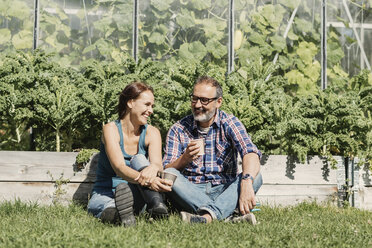 Happy farmers enjoying coffee break while sitting on grass against greenhouse - MASF03554