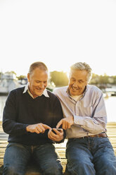 Glückliche ältere männliche Freunde, die ein Mobiltelefon auf einem Pier benutzen - MASF03560