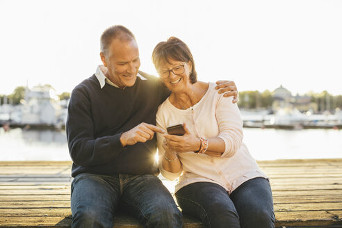 Glückliches älteres Paar, das auf dem Pier sitzend ein Mobiltelefon benutzt - MASF03561
