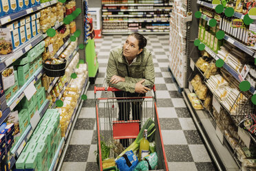 High angle view of thoughtful woman leaning on shopping cart at supermarket - MASF03596