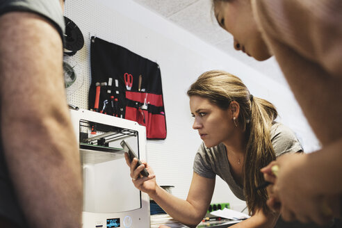 Eine Ingenieurin hält ein Smartphone in der Hand und betrachtet einen 3D-Drucker mit Kollegen - MASF03654