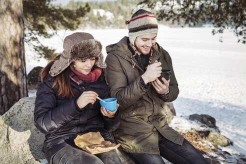 Couple having food while sitting on rock at field during winter - MASF03703