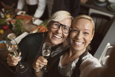 Cheerful mature women holding champagne flutes while taking selfie in kitchen stock photo