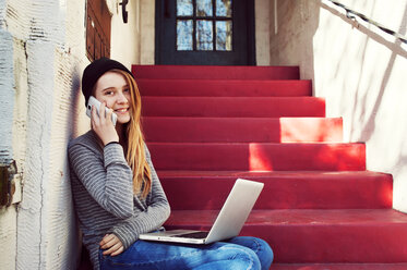 Porträt eines glücklichen Teenagers, der sein Smartphone bedient, während er auf den Stufen vor dem Haus sitzt - CAVF38018
