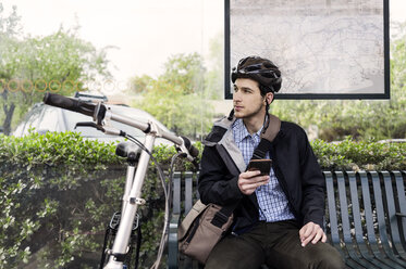 Junger männlicher Pendler, der an einer Bushaltestelle sitzt und sein Smartphone in der Hand hält - CAVF38036