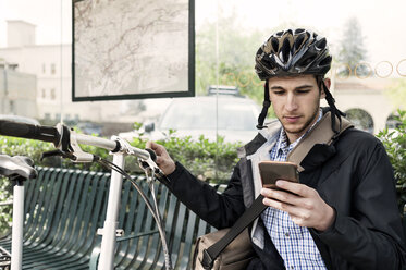 Männlicher Pendler, der an der Bushaltestelle sitzt und sein Smartphone benutzt - CAVF38037