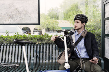 Nachdenklicher Geschäftsmann, der ein Fahrrad hält, während er an der Bushaltestelle sitzt - CAVF38038