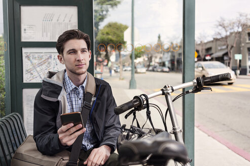 Nachdenklicher männlicher Pendler, der an der Bushaltestelle sitzt und sein Smartphone in der Hand hält - CAVF38039