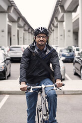 Porträt eines lächelnden Geschäftsmannes, der auf einem Fahrrad sitzt, mit Brücken und einem Parkplatz im Hintergrund - CAVF38046