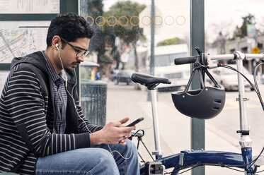Männlicher Pendler, der sein Smartphone benutzt, während er mit dem Fahrrad an der Bushaltestelle sitzt - CAVF38048