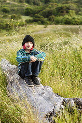 Porträt eines Jungen, der auf einem Baumstamm auf einem Feld sitzt und die Knie umarmt - CAVF38095