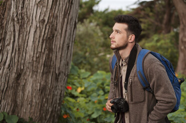 Male hiker looking away while holding camera in forest - CAVF38098