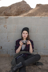 Portrait of confident tourist holding camera while sitting against retaining wall - CAVF38110