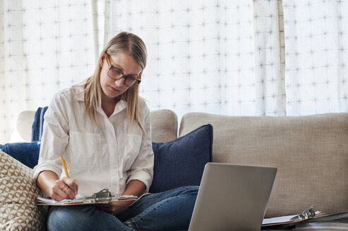 Frau schreibt auf einem Klemmbrett, während sie mit einem Laptop auf dem Sofa zu Hause sitzt - CAVF38135