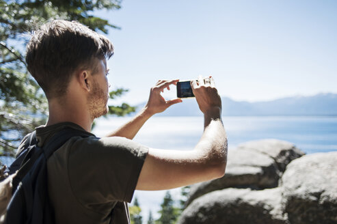 Rückansicht eines Mannes, der mit seinem Smartphone das Meer vor einem klaren blauen Himmel fotografiert - CAVF38156