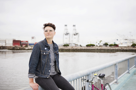 Frau mit Fahrrad schaut weg, während sie auf der Reling gegen das Meer sitzt, lizenzfreies Stockfoto