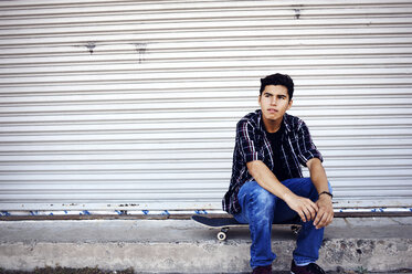 Thoughtful man looking away while sitting on skateboard against shutter - CAVF38185