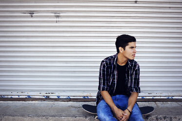 Man looking away sitting on skateboard against closed shutter - CAVF38187