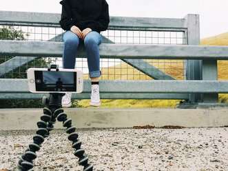 Nahaufnahme eines Mobiltelefons auf einem Stativ, das ein jugendliches Mädchen am Geländer sitzend fotografiert - CAVF38223