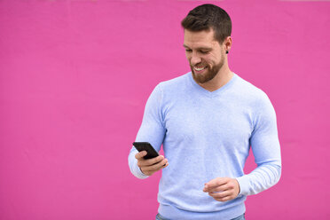 Lächelnder junger Mann schaut auf sein Smartphone, rosa Hintergrund - JSMF00143