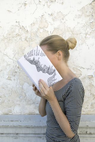 Frau bedeckt ihr Gesicht mit einem Buch und liest Gedichte vor einer Wand, lizenzfreies Stockfoto