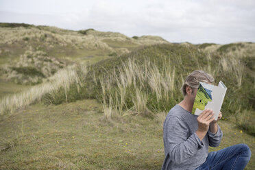 Man reading story book in the dunes, covering his face - PSTF00118