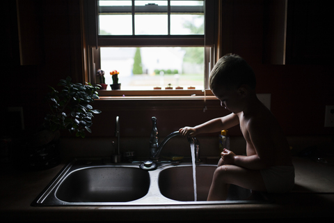 Verspielter Junge wäscht sich die Beine, während er an der Küchenspüle sitzt, lizenzfreies Stockfoto