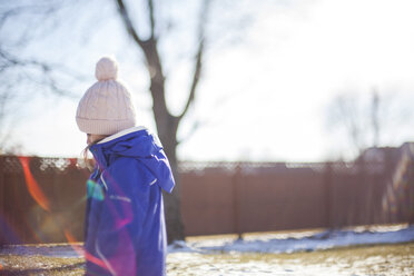 Side view of girl wearing knit hat standing in yard on sunny day during winter - CAVF38370