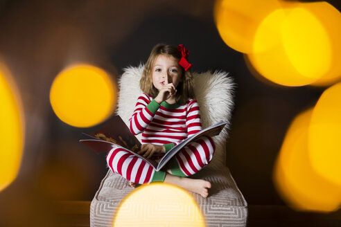 Porträt eines niedlichen Mädchens mit Finger auf den Lippen und einem Buch, das auf einem Stuhl zu Hause sitzt - CAVF38426