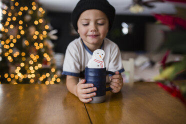 Süßer Junge hält Tasse mit Marshmallow Schneemann am Tisch in der Heimat - CAVF38430