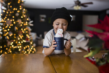 Glücklicher Junge hält Tasse mit Marshmallow-Schneemann am Tisch im Haus - CAVF38431