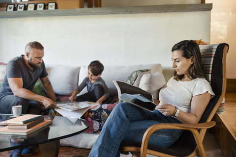 Frau liest eine Zeitschrift, während Vater und Sohn im Hintergrund zu Hause lernen, lizenzfreies Stockfoto