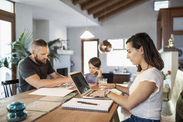 Woman working on laptop at dining table with family in background - MASF03763