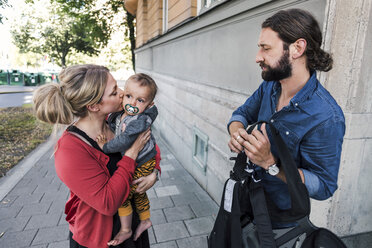 Berufstätige Mutter küsst ihren kleinen Jungen, während ein Mann auf dem Bürgersteig eine Trage hält - MASF03822