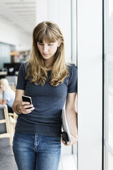 Junge Studentin benutzt ihr Smartphone, während sie sich in der Bibliothek ans Fenster lehnt - MASF03837