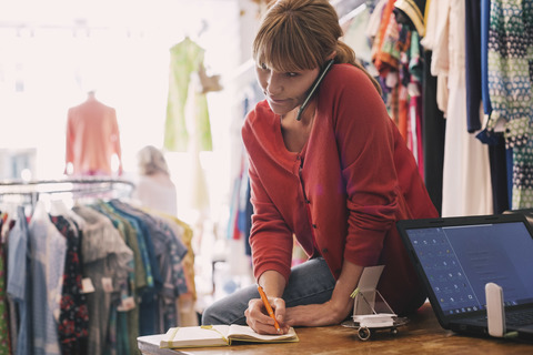 Frau schreibt Bestellung in Tagebuch, während sie in einem Secondhand-Laden mit ihrem Smartphone telefoniert, lizenzfreies Stockfoto