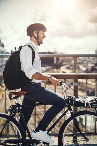 Lächelnder Geschäftsmann mit Fahrrad und Blick auf die Stadt, während er auf einer Brücke steht, lizenzfreies Stockfoto