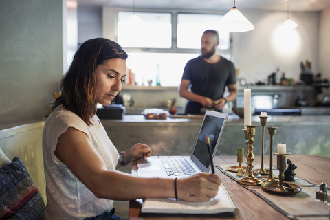 Seitenansicht einer Frau, die am Esstisch arbeitet, während ein Mann im Hintergrund steht, lizenzfreies Stockfoto