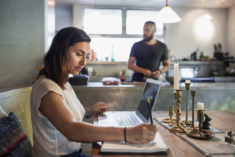 Side view of woman working at dining table while man standing in background stock photo