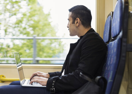 Seitenansicht eines jungen Mannes, der einen Laptop benutzt und durch ein Straßenbahnfenster schaut - MASF03920