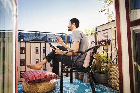 Side view of man holding smart phone and guidebook while looking away at balcony - MASF03923