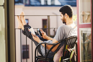 Seitenansicht eines Mannes, der ein Smartphone benutzt und einen Reiseführer auf einem Balkon hält - MASF03924