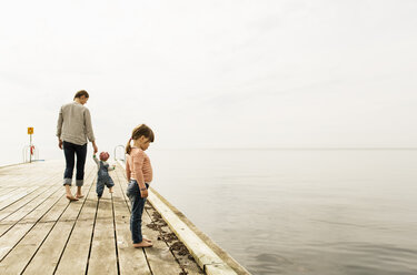 Side view of girl looking at sea while standing on pier with family walking in background against sky - MASF03926