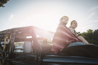 Side view of wonderlust couple wrapped in blanket sitting on jeep against sky - MASF03933