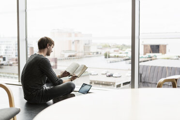 Seitenansicht eines Universitätsstudenten, der ein Buch liest, während er am Fenster in der Bibliothek auf dem Boden sitzt - MASF03936